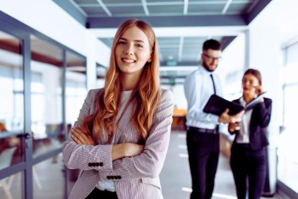 female standing in an office looking to camera, two colleagues talking in background